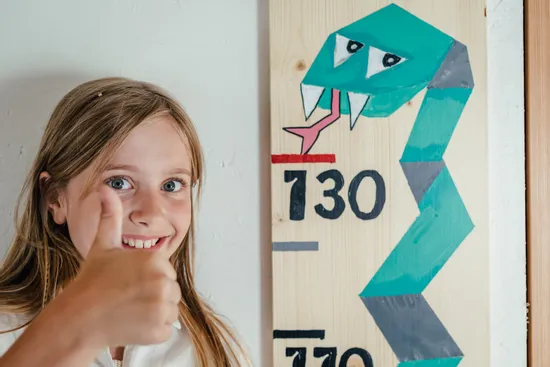 Lächelndes Mädchen zeigt Daumen hoch neben Größenmesslatte mit Schlangenmotiv