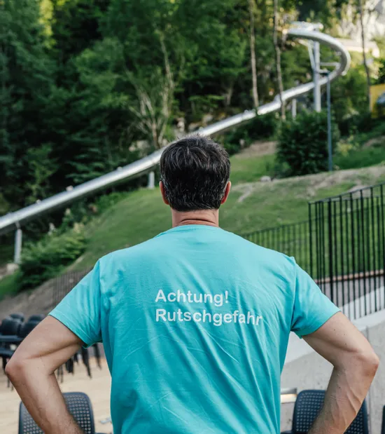 Mann im türkisfarbenen Shirt mit Aufdruck 'Achtung! Rutschgefahr' nahe einer Outdoor-Rutsche