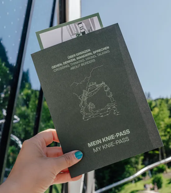 Hand hält ein schwarzes Heft mit der Aufschrift MEIN KNIE-PASS vor sonniger Berglandschaft
