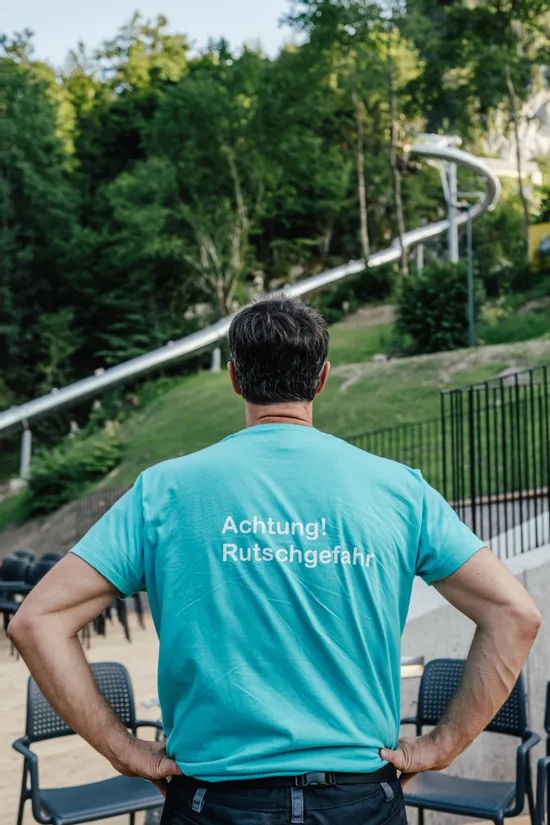 Mann im türkisfarbenen Shirt mit Aufdruck 'Achtung! Rutschgefahr' nahe einer Outdoor-Rutsche