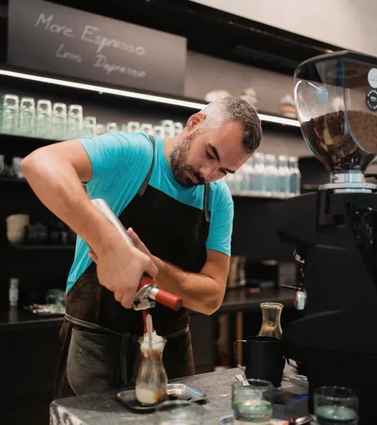 Barista mit blauem Shirt und Schürze bereitet Kaffee mit Sahne in modernem Café zu
