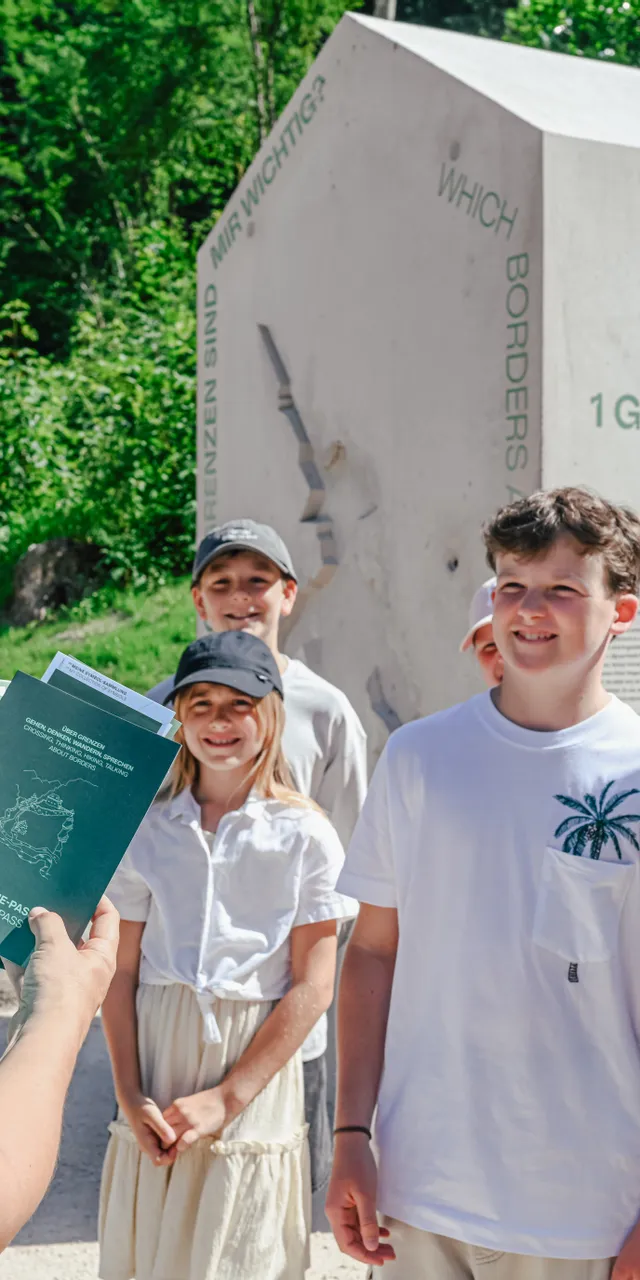 Guide mit Pässen spricht mit lachenden Kindern am Denkmal Grenzen und Übergänge in der Natur