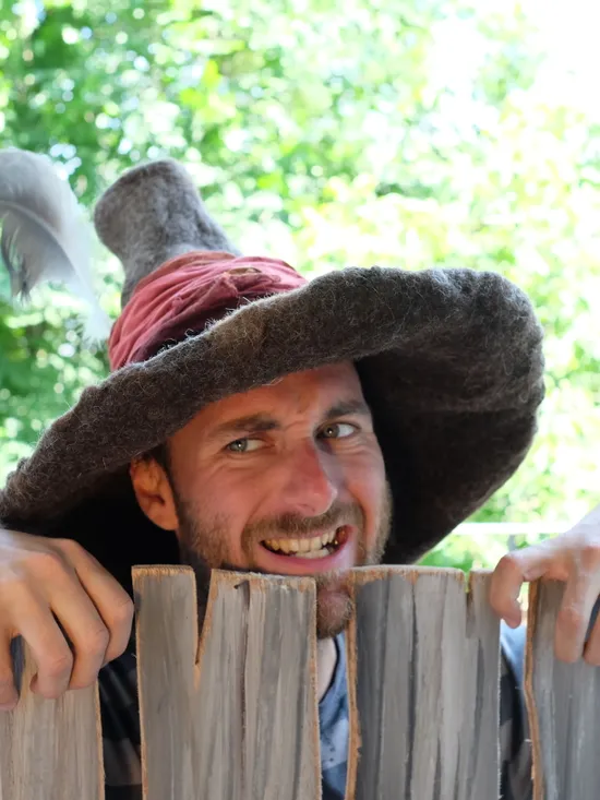 Mann mit fantasievollem Zaubererhut und Feder lugt über rustikalen Holzzaun