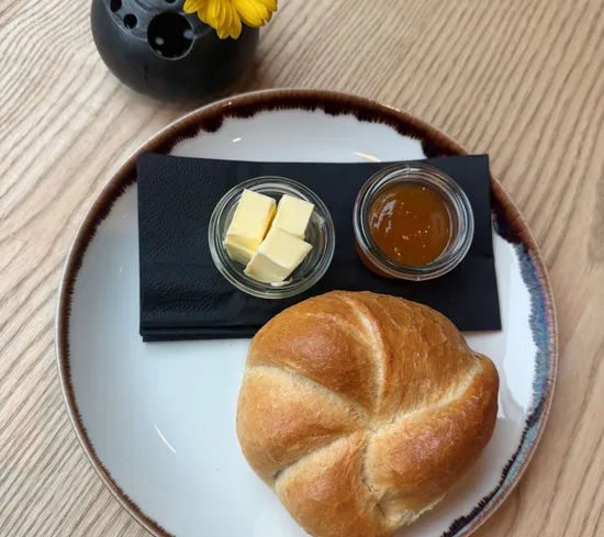 Frühstücksteller mit Brötchen, Butter, Marmelade neben gelber Blume in Vase