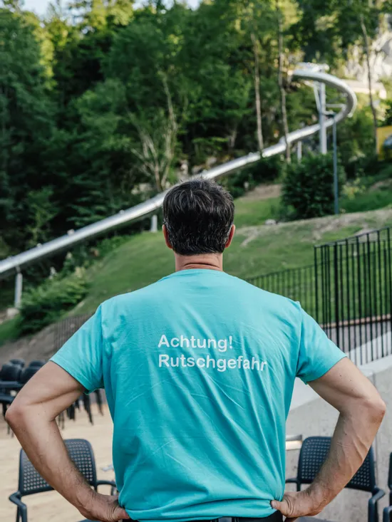 Mann im türkisfarbenen Shirt mit Aufdruck 'Achtung! Rutschgefahr' nahe einer Outdoor-Rutsche