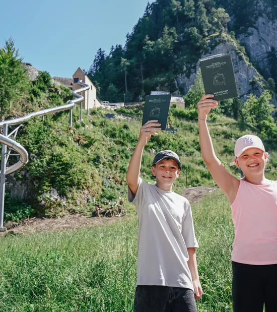 Drei lachende Kinder mit Wanderpässen vor Sommerrodelbahn in den Alpen