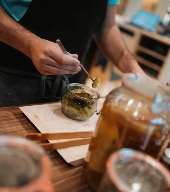 Person in apron using tweezers to handle pickled vegetables from glass jar