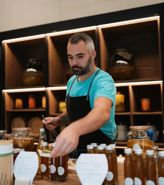 Man in apron offering honey samples at a market stall with glass bottles