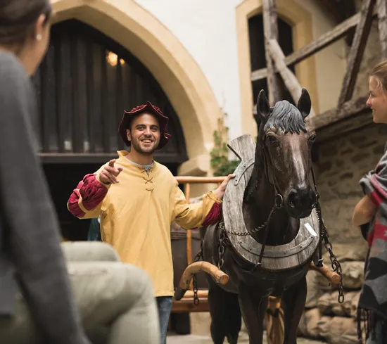 Medieval reenactor guiding visitors next to a wooden horse in a historic courtyard