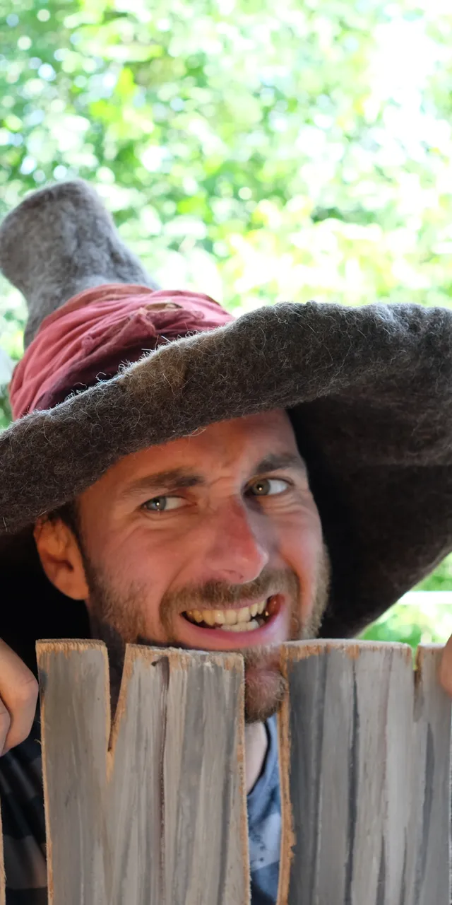 Mann mit fantasievollem Zaubererhut und Feder lugt über rustikalen Holzzaun