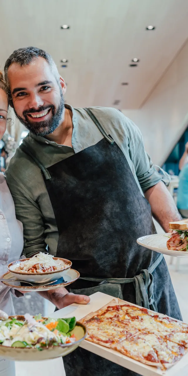 Smiling restaurant owners serving fresh pizza, salad, pasta, and sandwich in modern eatery
