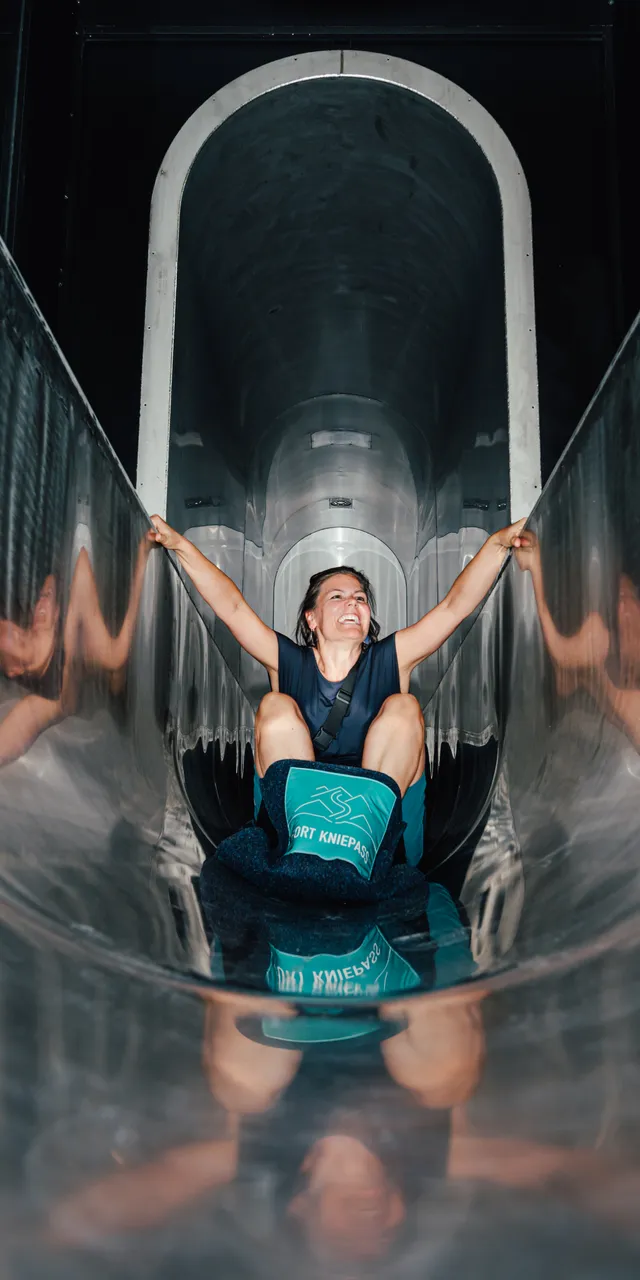 Smiling woman sliding down a shiny metal tunnel slide at an adventure park